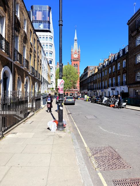 A wide city street in West Hampstead under clear blue skies, featuring residential buildings with brick facades and large windows on both sides. The sidewalk is clean with a concrete surface, and a black metal railing separates the pedestrian area from the road. Visible street furniture includes a streetlamp and a lamppost with a traffic sign attached. The street extends toward a prominent red-brick church with a tall spire and clock, serving as a central landmark in the background. Parked cars and mopeds line the street, while a few pedestrians walk along the pavement. Bright sunlight illuminates the scene, highlighting the pristine condition of the surroundings and emphasizing the neatness typical of well-maintained urban areas. This image reflects the tidy, organized atmosphere of West Hampstead, showcasing the importance of cleanliness and community upkeep, which a professional cleaning service such as Cleaners West Hampstead can assist with to avoid hidden cleaning charges in residential and commercial spaces.
