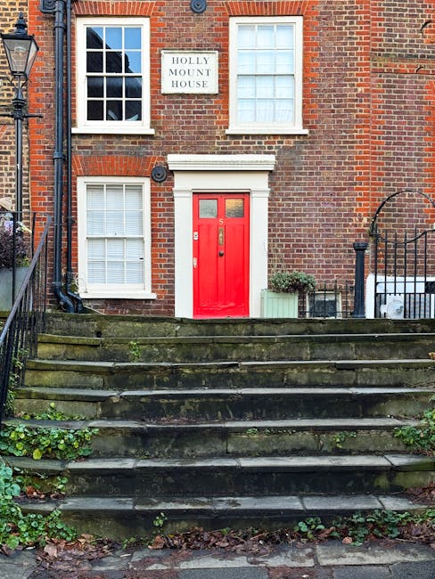 Exterior view of a historic brick building named Holly Mount House with a bright red front door and white window frames. The building features three visible windows, two on the first floor and one on the ground floor, all with white shutters. Stone steps with moss and small plants growing between them lead up to the entrance. A black wrought-iron railing runs along the right side of the steps. To the right of the door, there is a small green potted plant placed next to a window. A white sign with black text displays 'HOLLY MOUNT HOUSE' on the brick wall above the door. The image captures a well-maintained, classic urban residential façade, illustrating clean exterior surfaces suitable for professional cleaning or maintenance, reflective of services offered by Cleaners West Hampstead.
