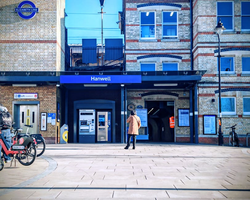 A wide city street in West Hampstead under clear blue skies, featuring residential buildings with brick facades and large windows on both sides. The sidewalk is clean with a concrete surface, and a black metal railing separates the pedestrian area from the road. Visible street furniture includes a streetlamp and a lamppost with a traffic sign attached. The street extends toward a prominent red-brick church with a tall spire and clock, serving as a central landmark in the background. Parked cars and mopeds line the street, while a few pedestrians walk along the pavement. Bright sunlight illuminates the scene, highlighting the pristine condition of the surroundings and emphasizing the neatness typical of well-maintained urban areas. This image reflects the tidy, organized atmosphere of West Hampstead, showcasing the importance of cleanliness and community upkeep, which a professional cleaning service such as Cleaners West Hampstead can assist with to avoid hidden cleaning charges in residential and commercial spaces.