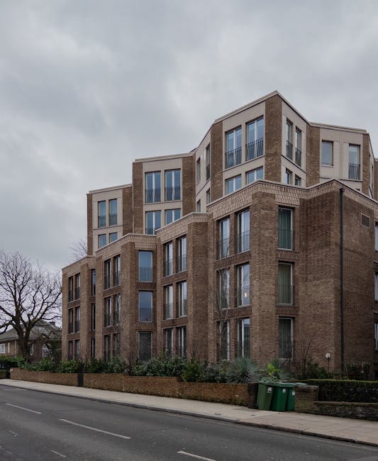 Photograph of a modern multi-story residential building featuring a combination of brick and light-colored paneling, with large glass windows and balconies. The building is situated along a paved street, with a low brick wall and greenery, including bushes and small trees, in the foreground. Overcast sky casts diffused light over the scene. The image highlights the clean exterior surfaces of the building, consistent with professional deep cleaning and maintenance standards, as provided by Cleaners West Hampstead for end of tenancy cleaning in West Hampstead.