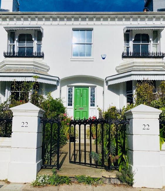 Image of a white residential building with a bright green front door situated behind a black wrought iron gate. The building features decorative railings on the small balconies above the entrance, with large, clear windows on the upper floors. The front garden is landscaped with lush, green plants and flowers, adding to the tidy appearance. The gate and pathway leading to the door are well-maintained, and daylight illuminates the scene, emphasizing the clean and pristine condition of the exterior. This setting exemplifies professional surface cleaning and maintenance, consistent with the services offered by Cleaners West Hampstead for deep cleaning and sanitisation of domestic properties. The image underscores the presentation of a well-kept home, ready for tenancy or resale.