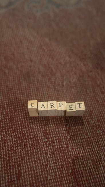 Close-up image of a textured carpet with a reddish-brown hue, featuring small, evenly spaced loops. In the center, the words 'CARPET' are spelled out with small, cream-colored wooden letter blocks arranged on the surface. The lighting is soft and even, highlighting the cleanliness and neat presentation of the area. This image reflects the focus on surface cleaning and maintenance typical of residential or commercial carpet cleaning services offered by Cleaners West Hampstead, situated on Mill Lane in West Hampstead NW6, as described on their website.