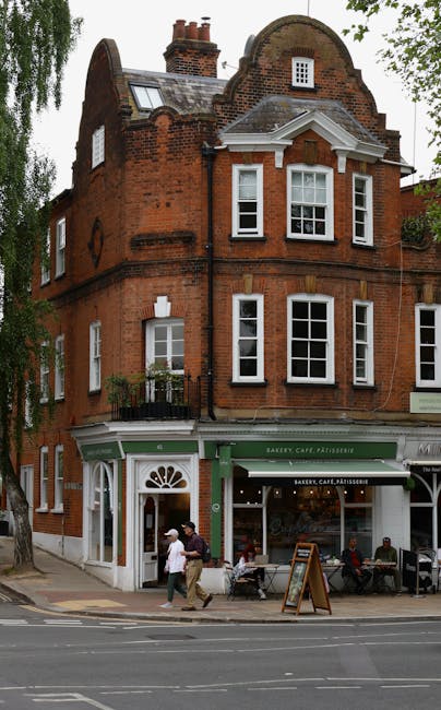 The image shows a multi-story red brick building with a ground-floor bakery, cafe, and pâtisserie, featuring large glass windows and a green awning. The upper floors have several white-framed sash windows, some with decorative stone lintels and a small balcony with black wrought iron railings on the second floor. The building has a curved gable roof with brick chimneys and rooftop dormer windows. Outside, three pedestrians are walking past the storefront, which displays outdoor seating and a chalkboard sign. The scene is illuminated by natural daylight, with surrounding trees partially visible on the left, indicating an urban street setting. This image represents a typical commercial property in West Hampstead, maintained with clean and inviting facades, accentuating the area's mixed residential and retail character, suitable for promoting professional surface cleaning and maintenance services in the vicinity, as offered by Cleaners West Hampstead.