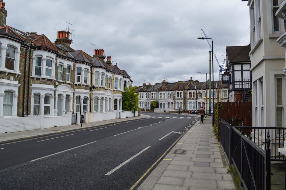 A row of elegant Victorian-style residential buildings along West End Lane in West Hampstead, featuring yellow and brown brick facades with white window trims. The street is lined with evenly spaced, leafless trees and classic black lampposts. A sidewalk with a brick and wrought-iron fence surrounds the front gardens, which contain neatly maintained plants and flowering trees with white blossoms. The sky above is clear blue with some scattered clouds, casting bright sunlight over the scene, highlighting the historic architecture and well-kept exterior surroundings. This image represents a peaceful, suburban neighborhood viewed from street level, emphasizing the picturesque and tidy environment.